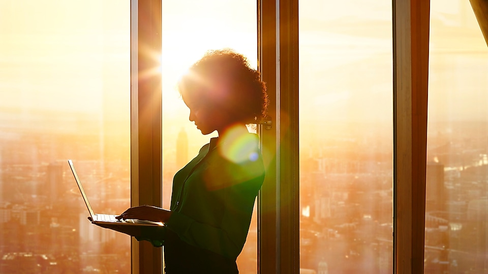 Mujer parada usando una laptop contra una puesta de sol detrás de ventanas en un edificio