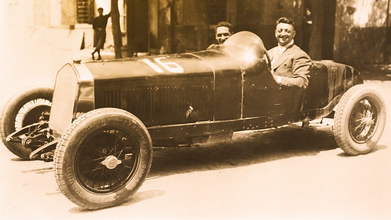 Fotografía en blanco y negro de Enzo Ferrari sentado en uno de sus primeros autos Ferrari