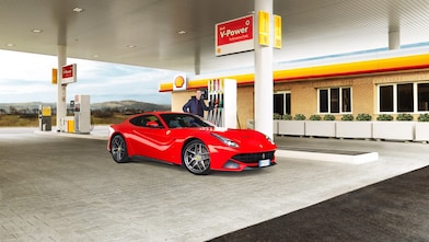 Ferrari rojo en una estación de servicio Shell con un hombre apoyado sobre un surtidor