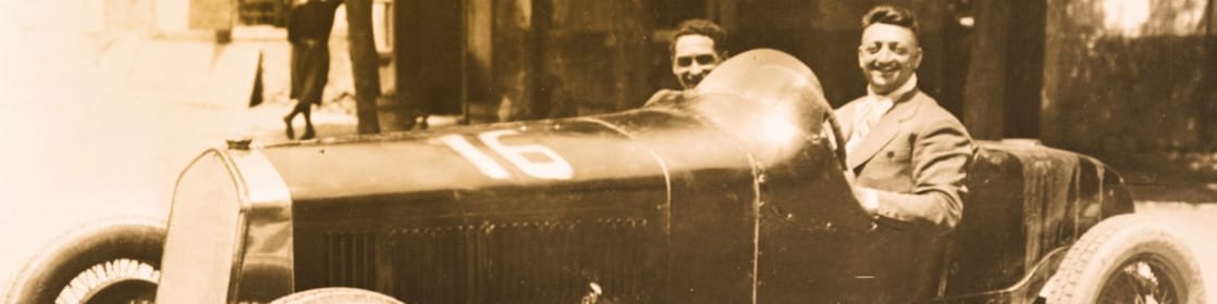 Fotografía en blanco y negro de Enzo Ferrari sentado en uno de sus primeros autos Ferrari