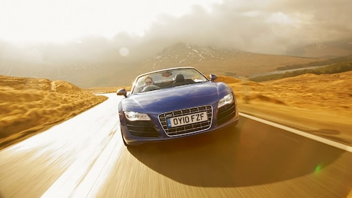 Auto azul descapotable, de cara, circulando por la campiña rodeado de paisajes montañosos