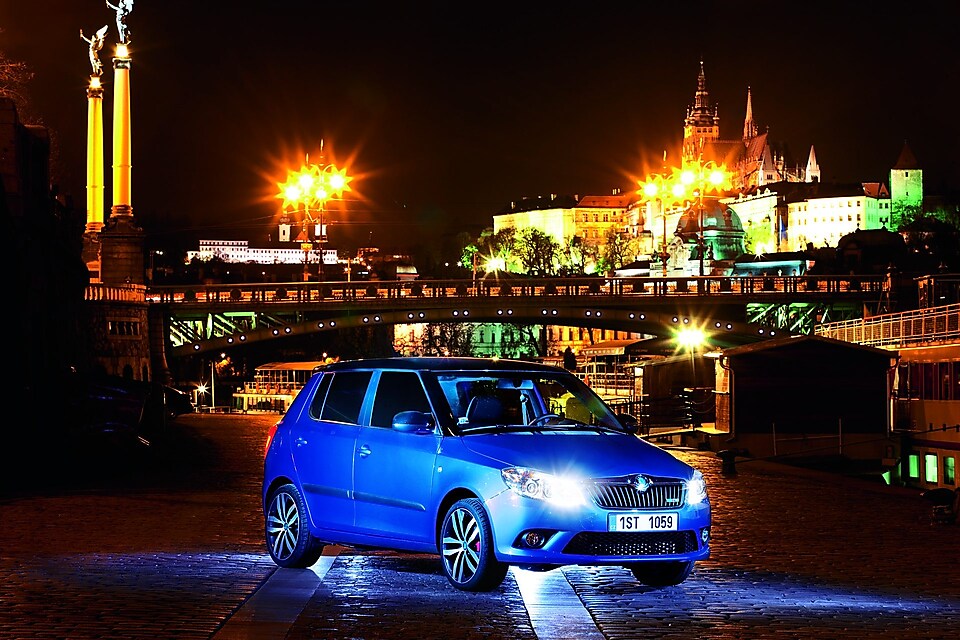 Auto azul estacionado en una calle de Praga con un puente y el castillo al fondo, de noche