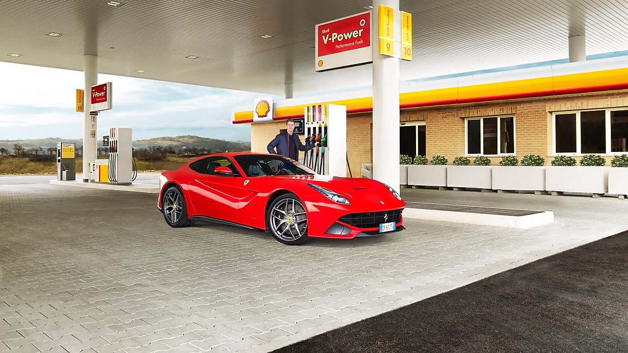 Ferrari rojo en una estación de servicio Shell con un hombre apoyado sobre un surtidor