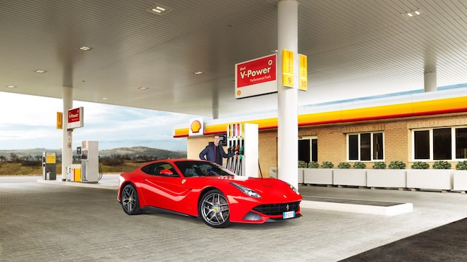 Ferrari rojo en una estación de servicio Shell con un hombre apoyado sobre un surtidor