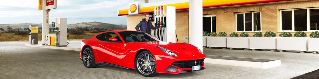 Ferrari rojo en una estación de servicio Shell con un hombre apoyado sobre un surtidor